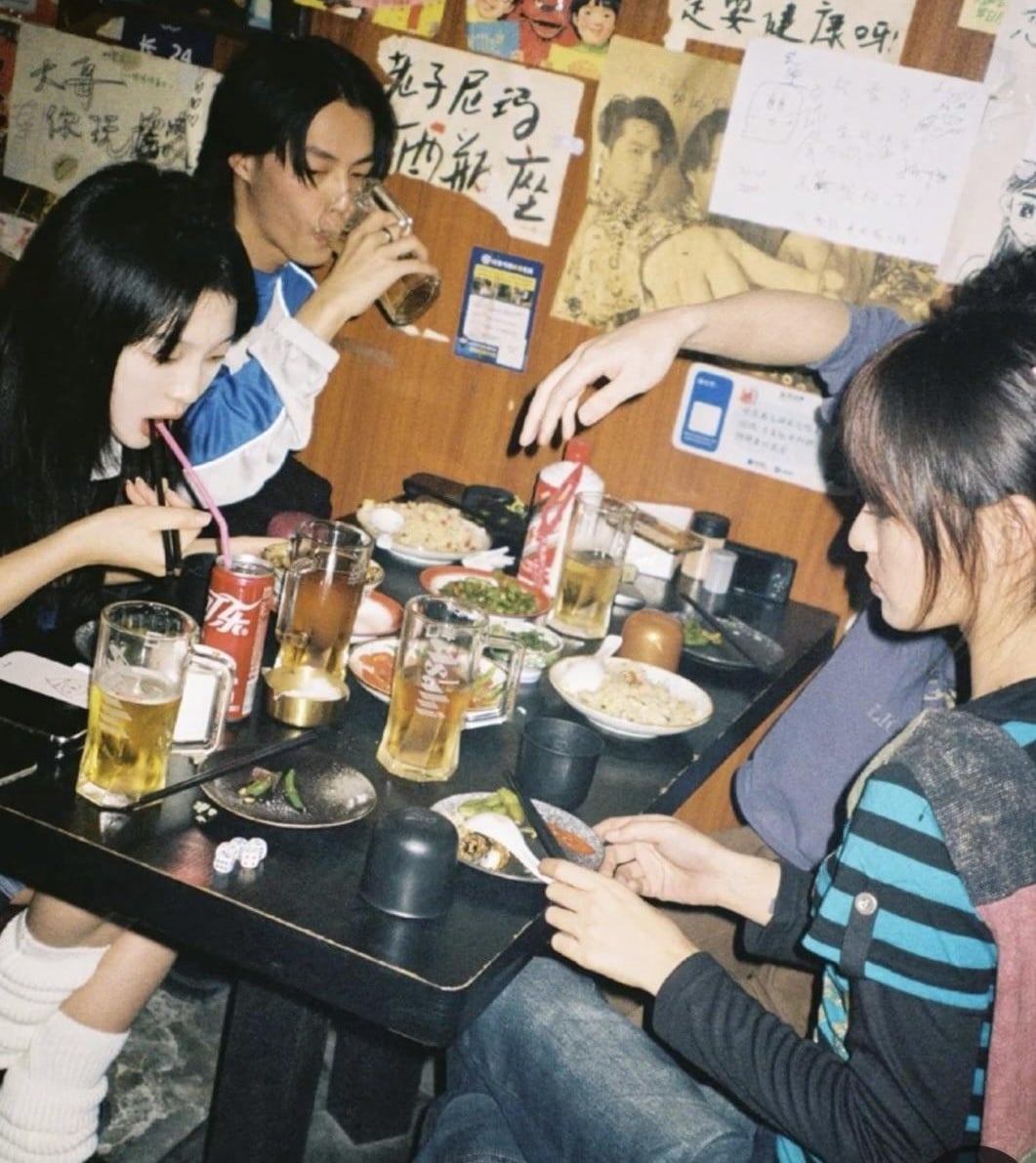 Three people sit at a restaurant table with dishes, mugs of beer, and soda cans, eating and drinking. One person pours a drink while others use chopsticks and a fork. The walls are covered with handwritten notes and posters.