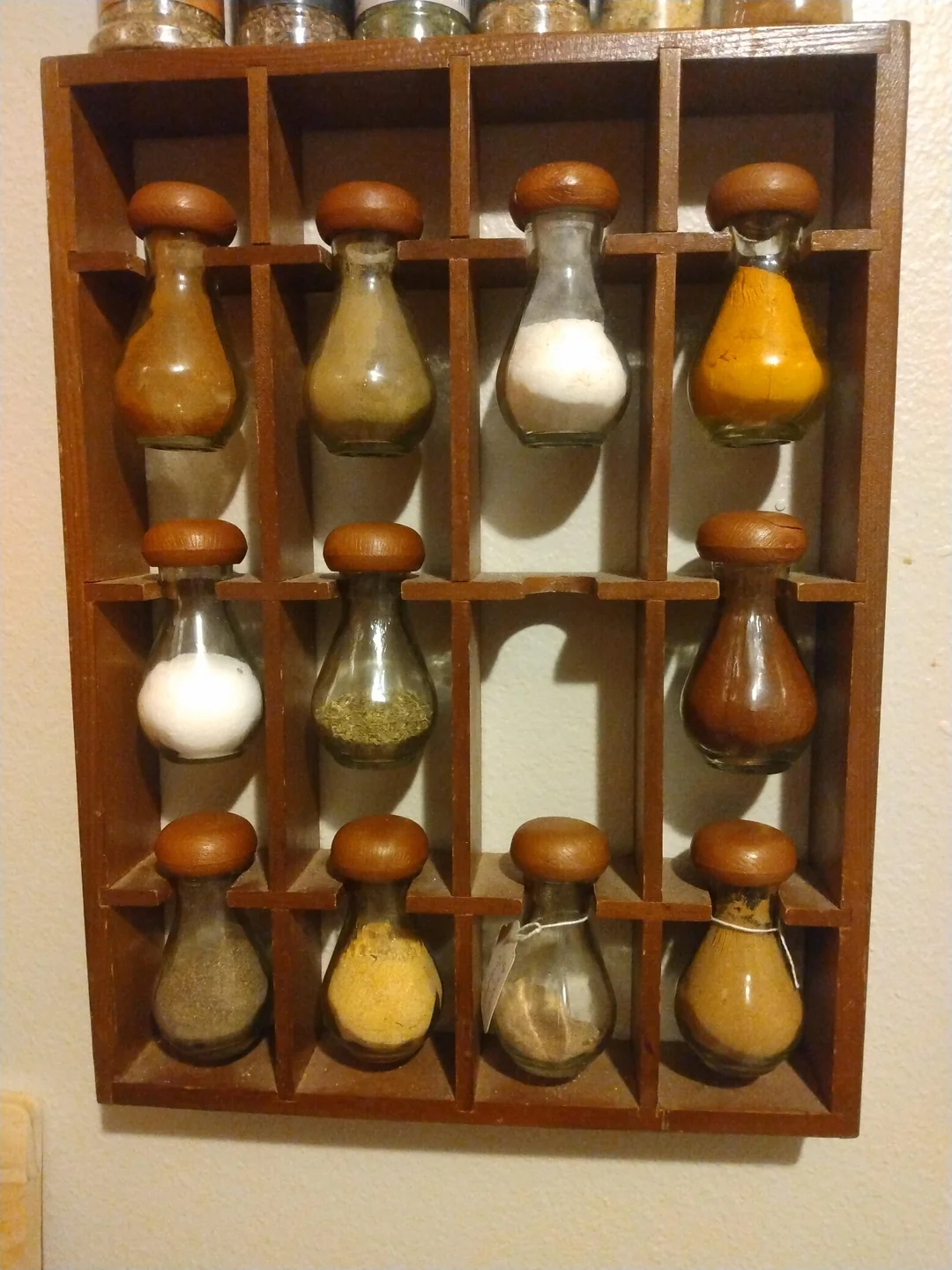 A wooden spice rack with 12 glass jars, each filled with different spices or seasonings. The jars have round brown caps and are neatly arranged in a 3 by 4 grid on a light-colored wall.
