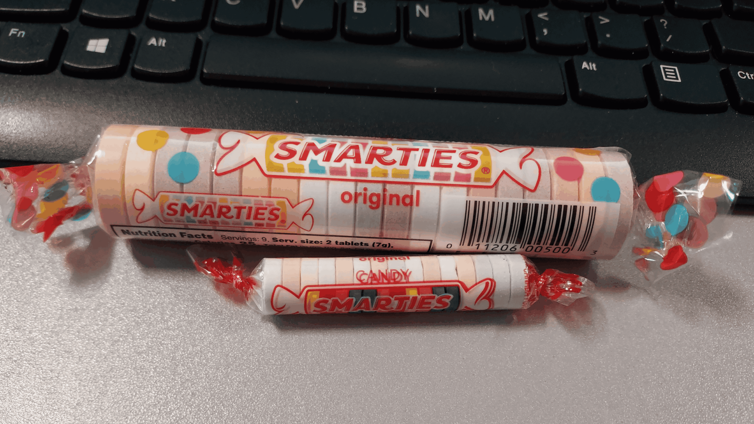 Two rolls of Smarties candy—one large, one small—sit on a desk in front of a computer keyboard.