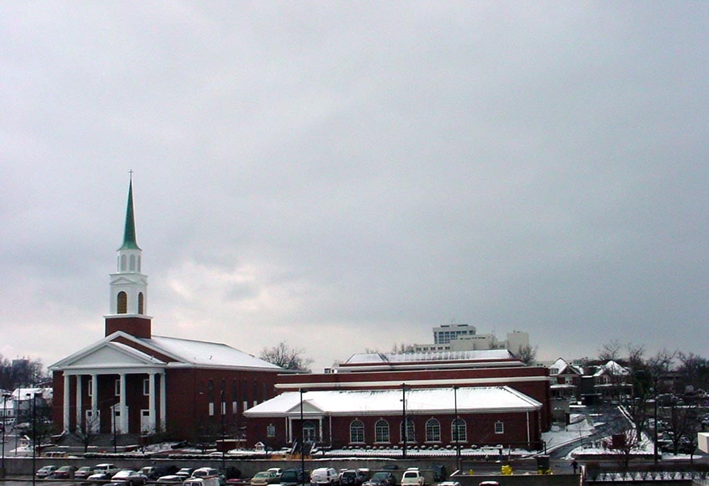A red-brick church with a tall white steeple and a green roof stands beside other buildings. Snow covers the ground and rooftops. Cars are parked in front; the sky is overcast.