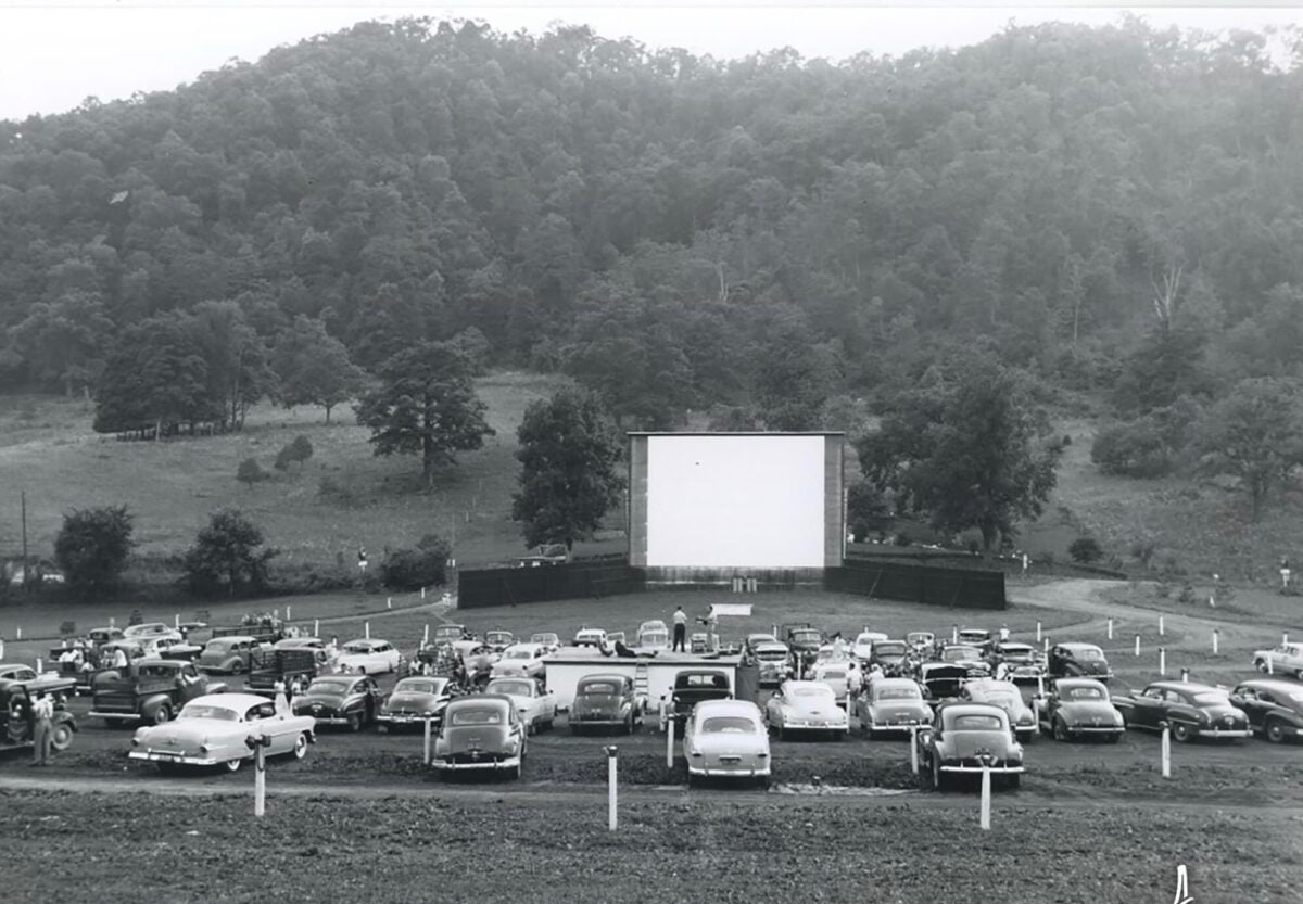 Black-and-white photo of a drive-in theater with vintage cars parked facing a large blank screen, surrounded by grassy hills and trees in the background.
