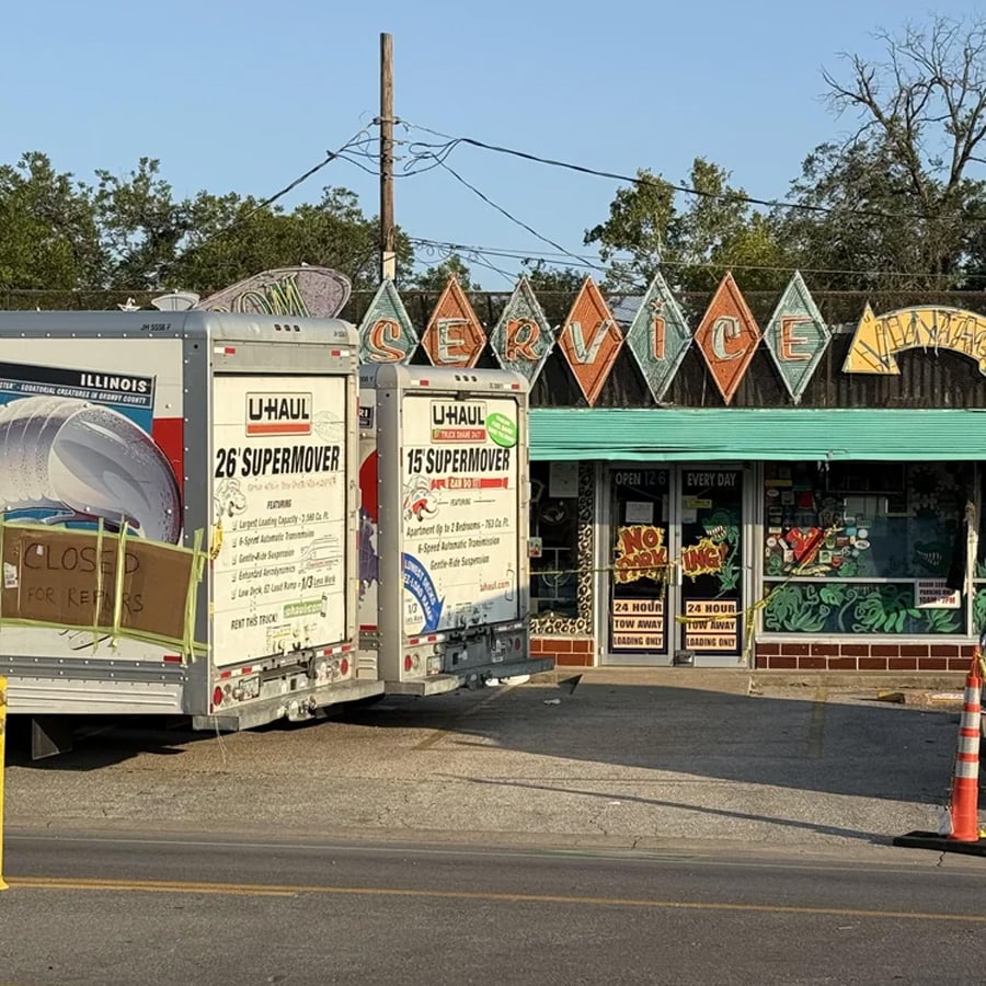 Two U-Haul trucks are parked in front of a retro-style diner with a colorful “Service” sign. The diner windows display various signs and the building has a teal roof. Trees and power lines are visible in the background.