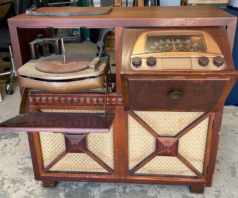 A vintage wooden console with a tilt-out turntable, an old radio dial with knobs, a pull-out shelf, and speaker panels below, sitting on a worn floor.