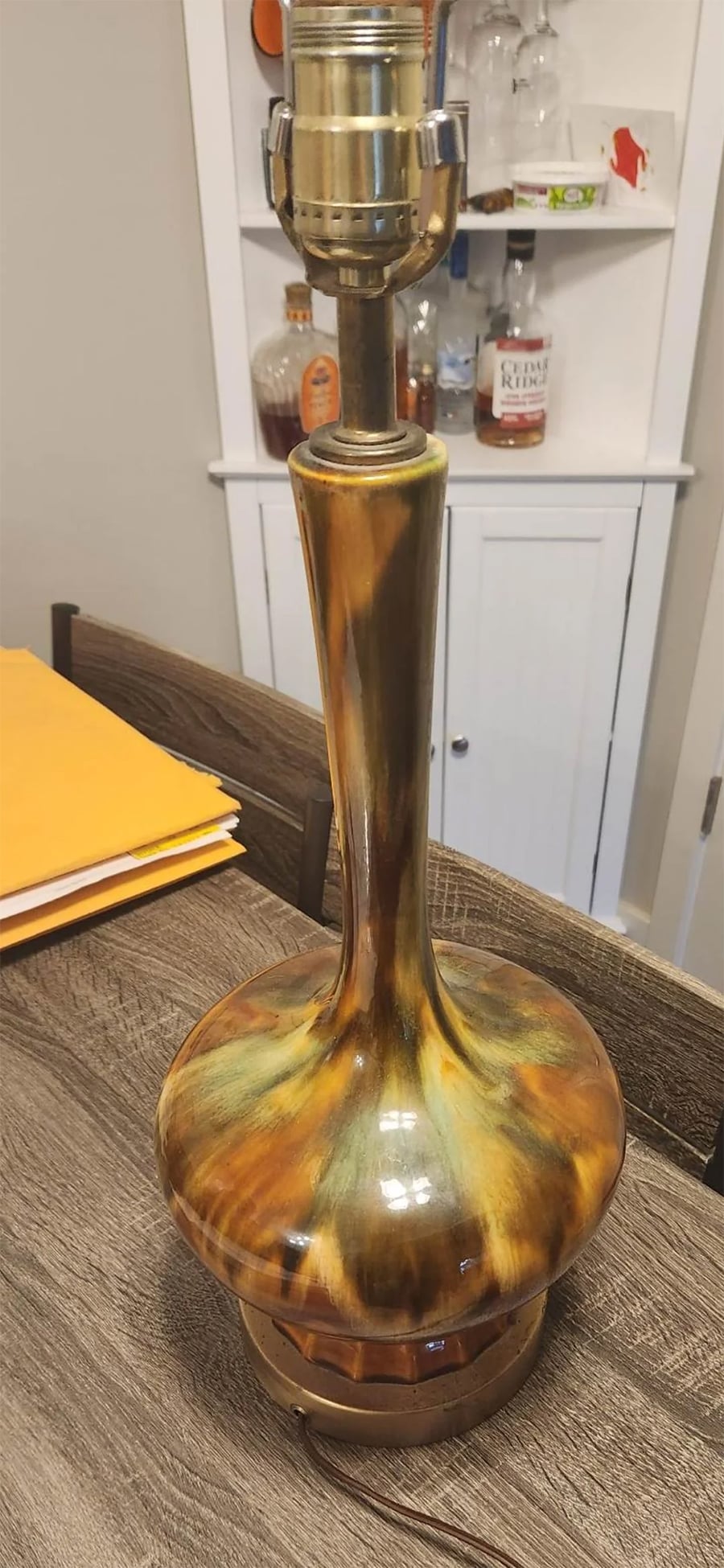 A vintage lamp with a bulbous, multicolored base sits on a wooden table. Papers and bottles are visible in the background.