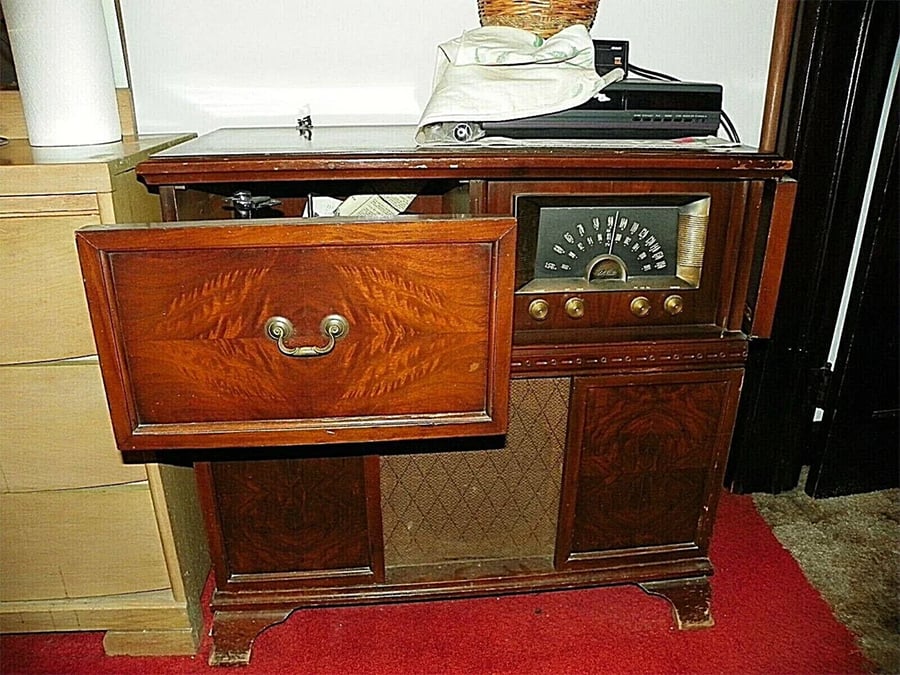 A vintage wooden radio and record player cabinet with an open drawer, showing old radio dials and a woven speaker cover, on a red carpet in a room.