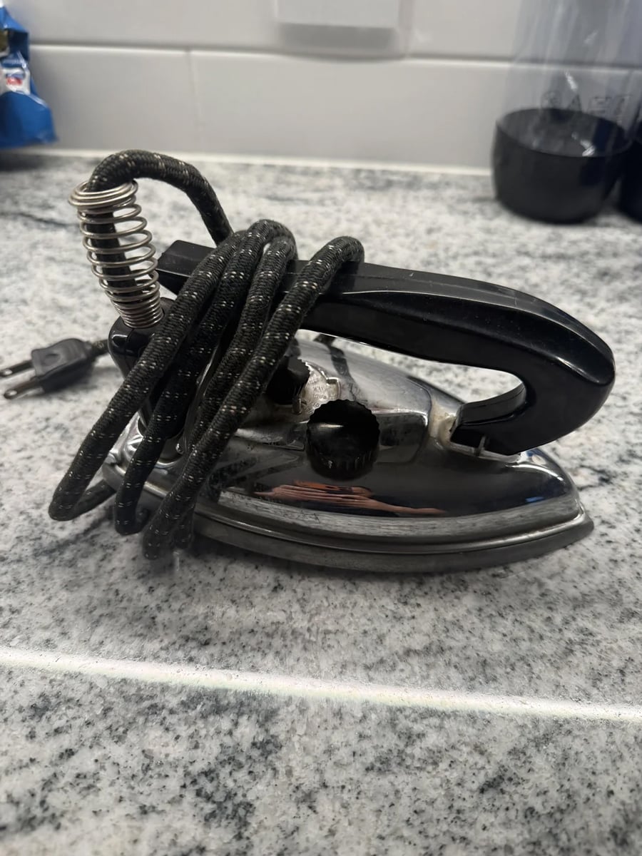 A small vintage metal iron with a black handle and coiled cord wrapped around it, sitting on a gray speckled countertop.