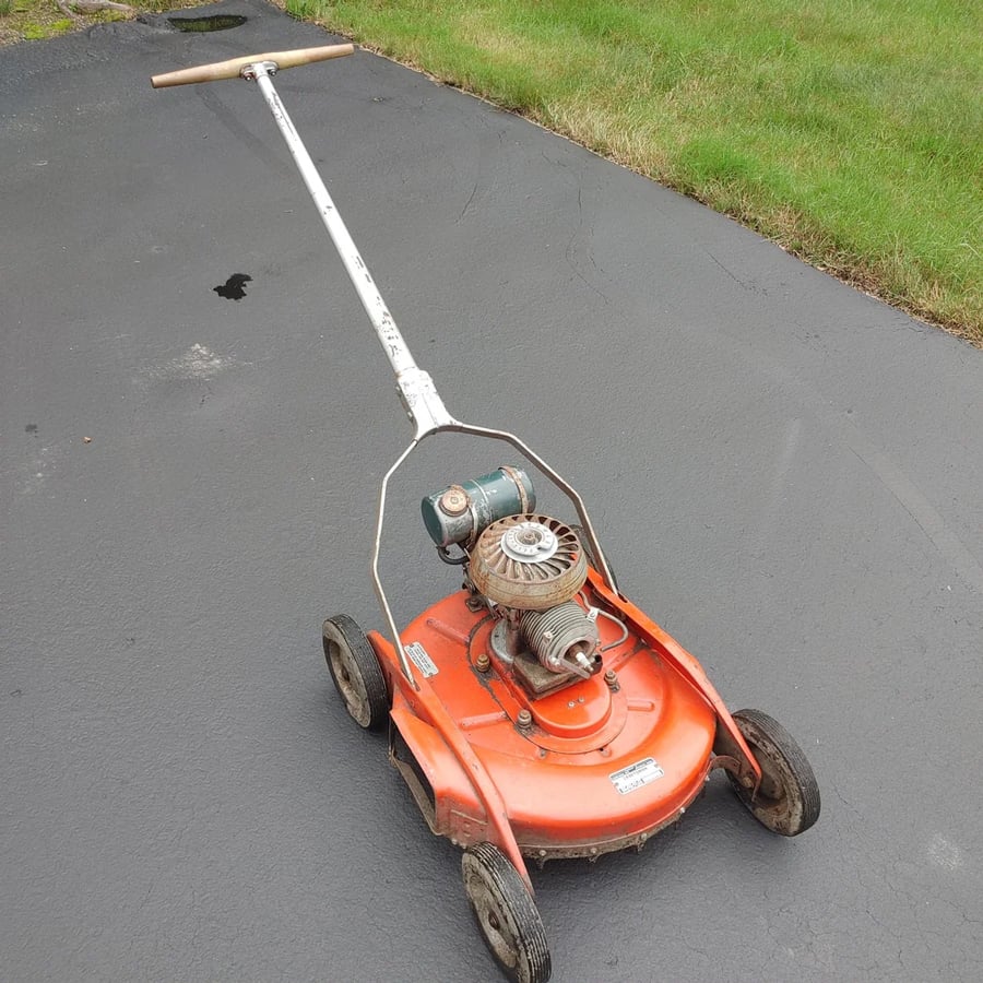 An old-fashioned, orange push lawn mower with four wheels and an exposed engine is on a paved driveway next to a patch of grass.