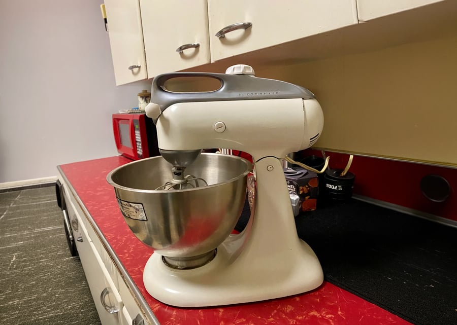 A white stand mixer with a stainless steel bowl sits on a red kitchen countertop. White cabinets and a red microwave are visible in the background.