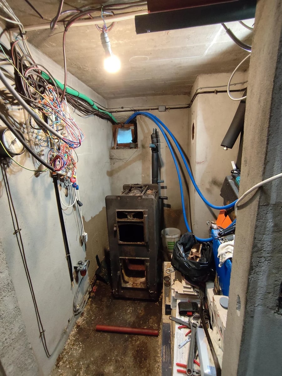 A small, cluttered basement room with exposed wiring, a metal stove, blue pipes, and various tools and materials scattered on shelves and the floor. A single light bulb hangs from the ceiling.