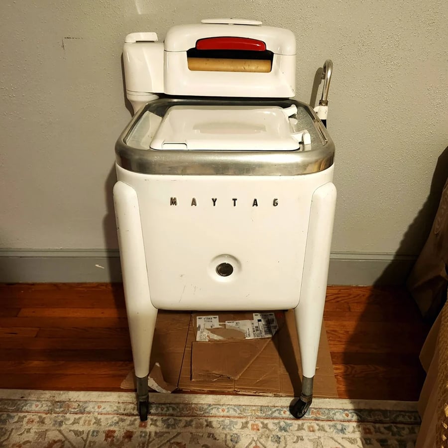 Vintage Maytag washing machine with wringer on top, white body, metal trim, and wheels, positioned against a beige wall on a wooden floor, with a cardboard box stored underneath.