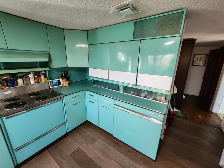 A retro kitchen with teal cabinets, a matching dishwasher, an electric stove, and various kitchen items on the counter. The floor is wooden, and the style appears vintage, likely from the mid-20th century.