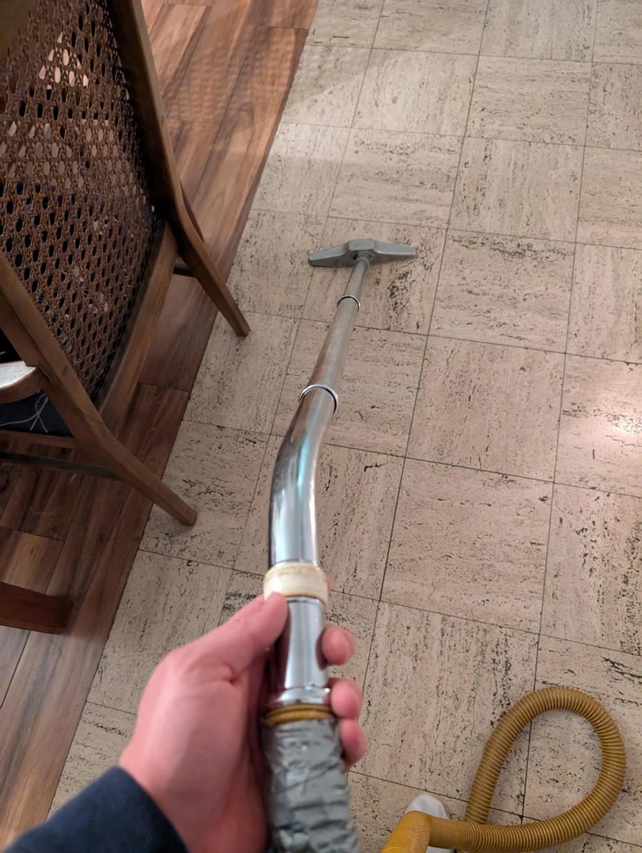 A hand holding a vacuum cleaner hose, cleaning a tiled floor next to a wooden chair. Part of the vacuum’s yellow cord is visible on the floor.
