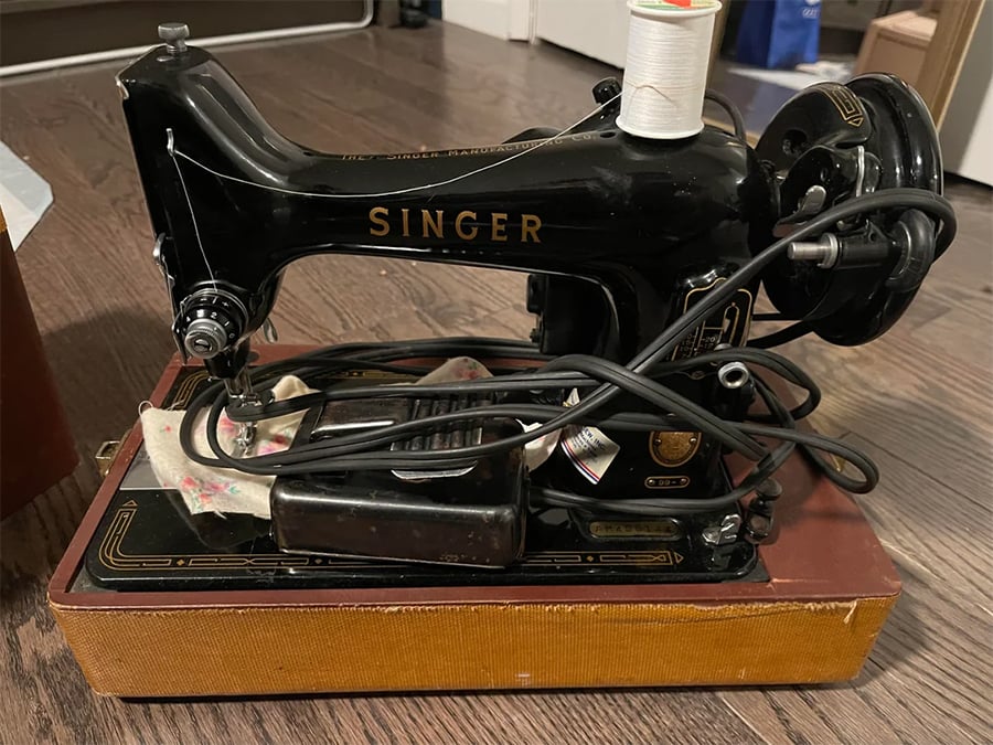 A vintage black Singer sewing machine with a spool of white thread on top, sitting on a brown base, electric cords and pedal draped over it, placed on a wooden floor.