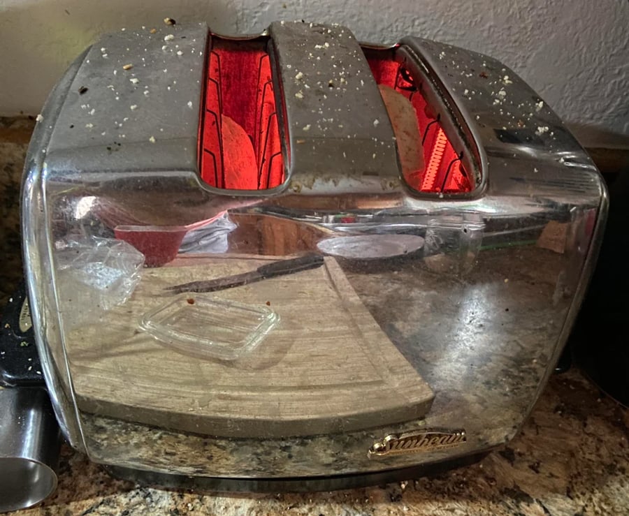 A dirty stainless steel toaster with crumbs on top is toasting bread. The toaster reflects a cluttered kitchen counter with a cutting board and dish.
