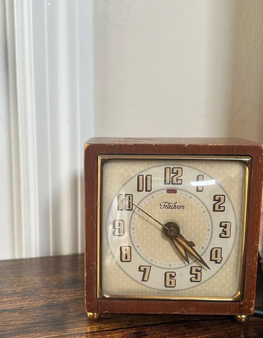 A vintage Telechron alarm clock with a wooden frame sits on a wooden surface. The clock shows the time as 4:28. The background features a white wall and a vertical molding strip.