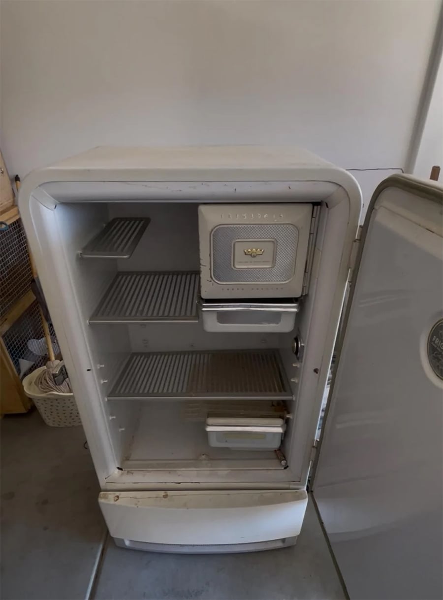 An open vintage white refrigerator with empty shelves and drawers, showing some rust and wear. The freezer compartment is closed, and the fridge door is fully open.