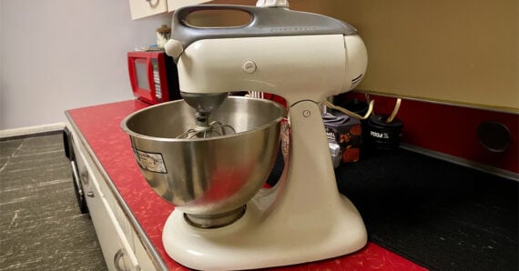 A white stand mixer with a metal bowl sits on a red countertop in a kitchen, with a red microwave and various containers in the background.