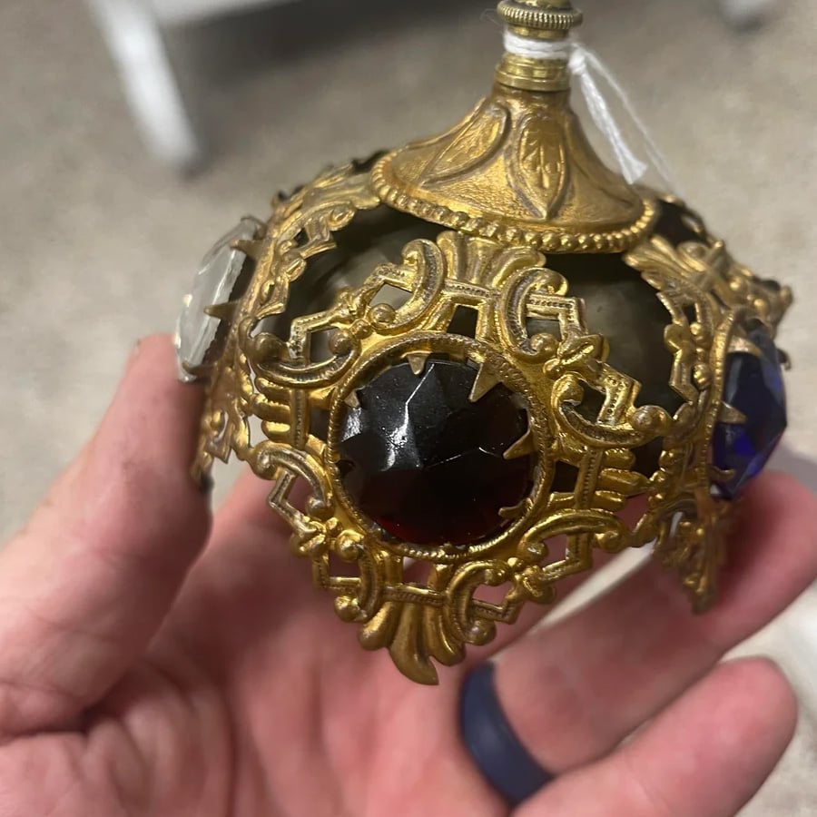 A hand holds an ornate, gold-colored metal object with intricate openwork and large, colored glass gems set in its sides, resembling a vintage decorative ornament or finial.