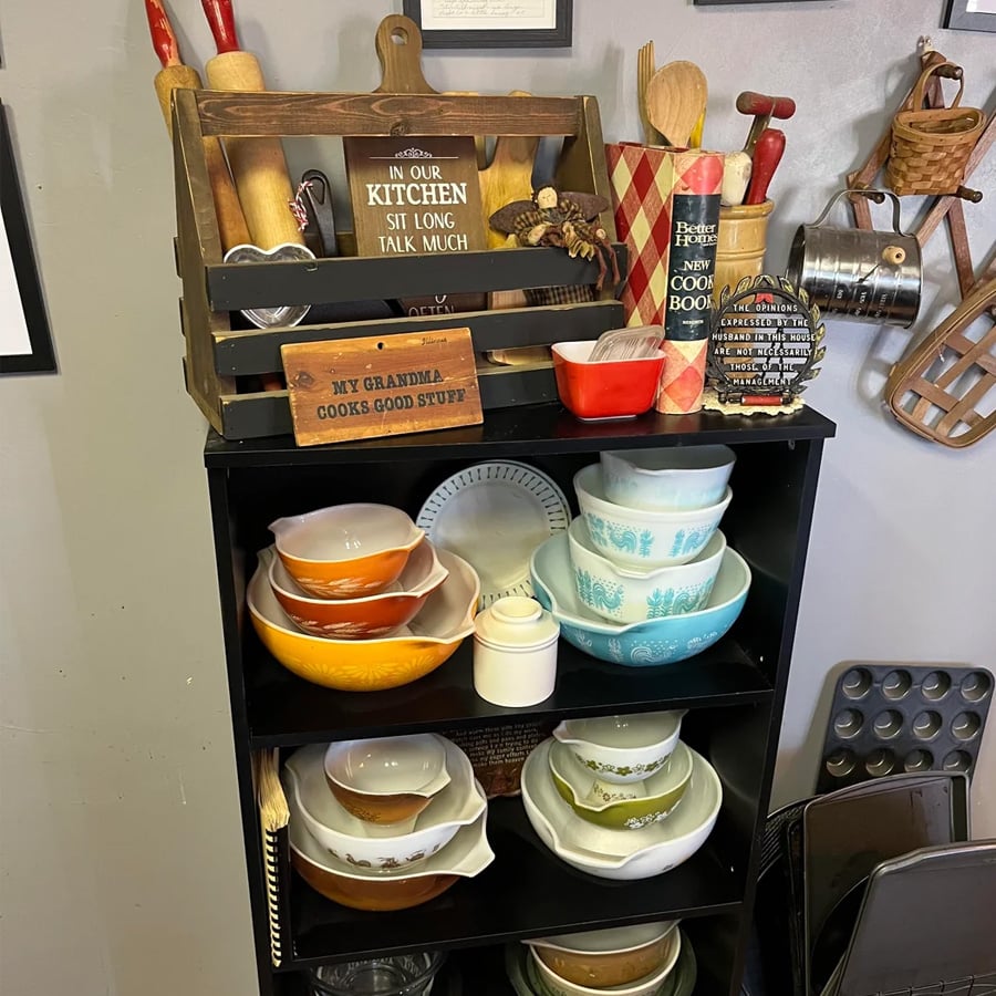 A black shelf holds vintage mixing bowls, dishes, cookbooks, and kitchen utensils. Signs say “IN OUR KITCHEN SIT LONG TALK MUCH” and “MY GRANDMA COOKS GOOD STUFF.” Various cookware is on the wall and surrounding the shelf.
