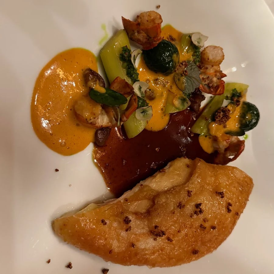 A plated dish featuring a golden-brown pastry slice, colorful vegetables, and lobster pieces, garnished with green herbs and served with two different sauces on a white plate.