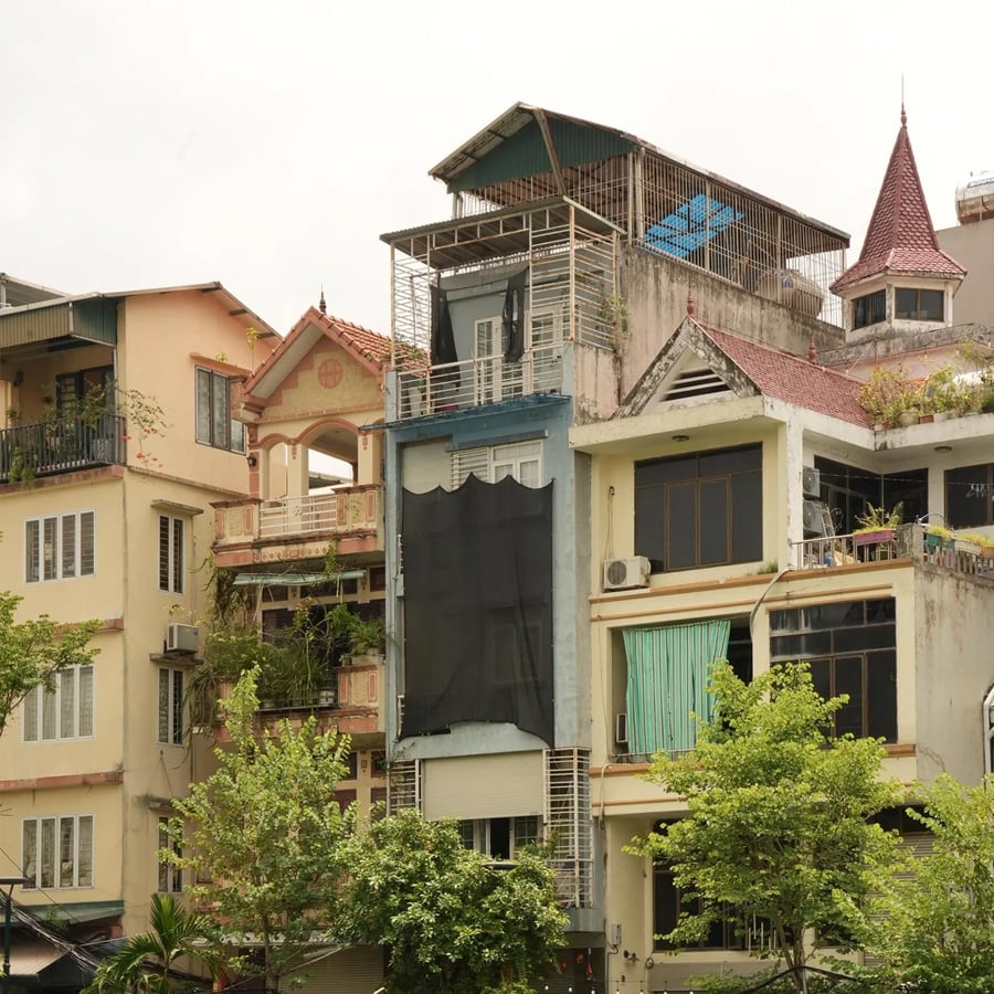 A group of narrow, multi-story buildings with balconies, rooftop structures, plants, and laundry hanging outside, surrounded by green trees under a cloudy sky.