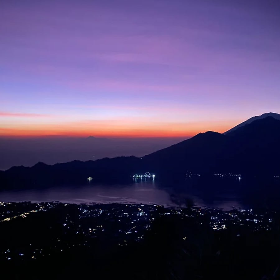 A vibrant sunset with purple and orange hues over distant mountains, a calm lake, and a town below with scattered lights twinkling in the evening.