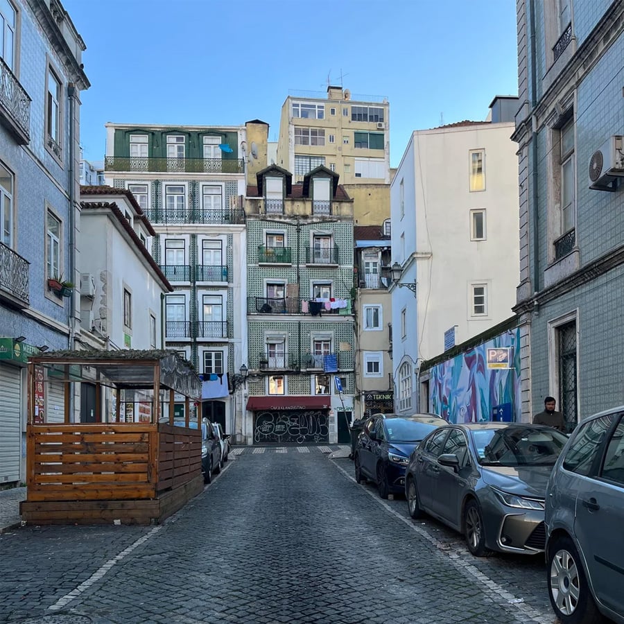 A narrow cobblestone street lined with parked cars, colorful apartment buildings, balconies with laundry, and a wooden structure on the left, under a clear blue sky.