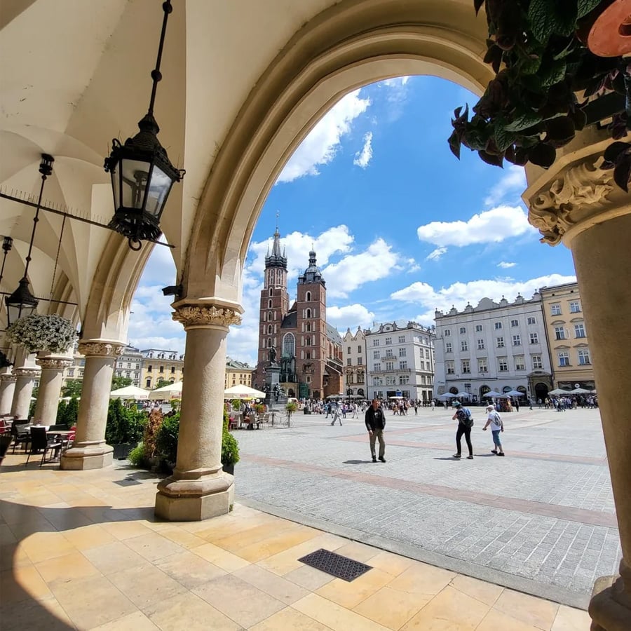 View of a historic city square through stone arches, with people walking, outdoor seating, classic buildings, and a tall church under a partly cloudy sky.