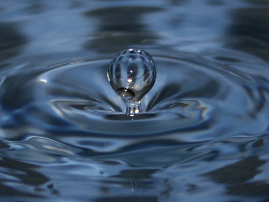 A close-up of a single water droplet rising above the surface, creating ripples in a calm, blue liquid.