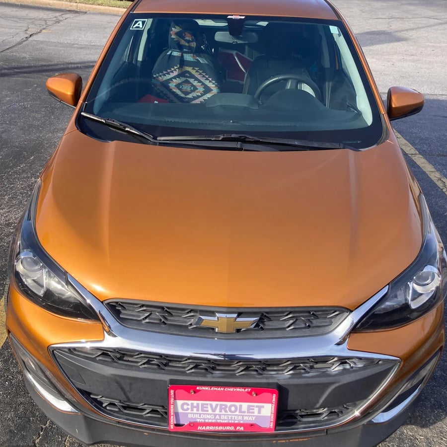 An orange Chevrolet car with a decorative patterned seat cover is parked in a lot. The windshield reflects the sky, and the front license plate holder shows a pink Chevrolet dealership tag.