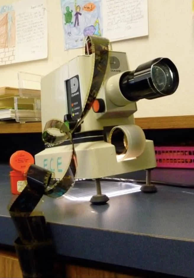A film strip is draped over a vintage slide projector on a table, with children’s drawings hanging on the wall in the background.