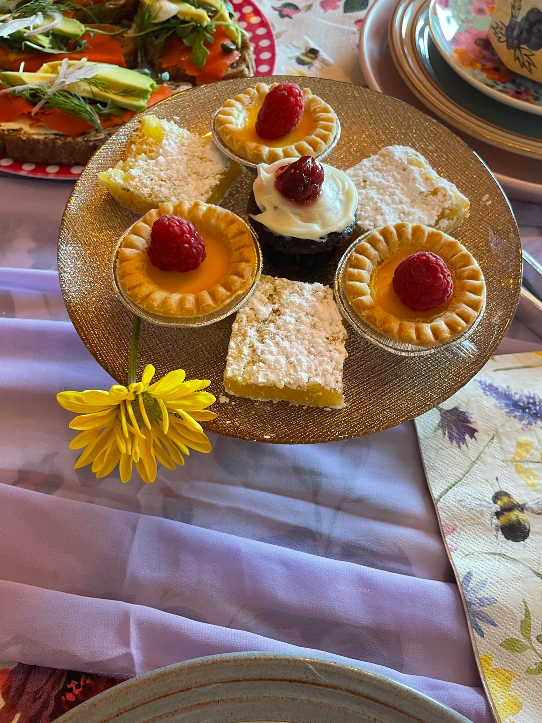 A gold plate holds tarts with raspberries, a cupcake with cream and berry, and a powdered sugar bar. A yellow flower rests on the plate, with a salad and teacup in the background.