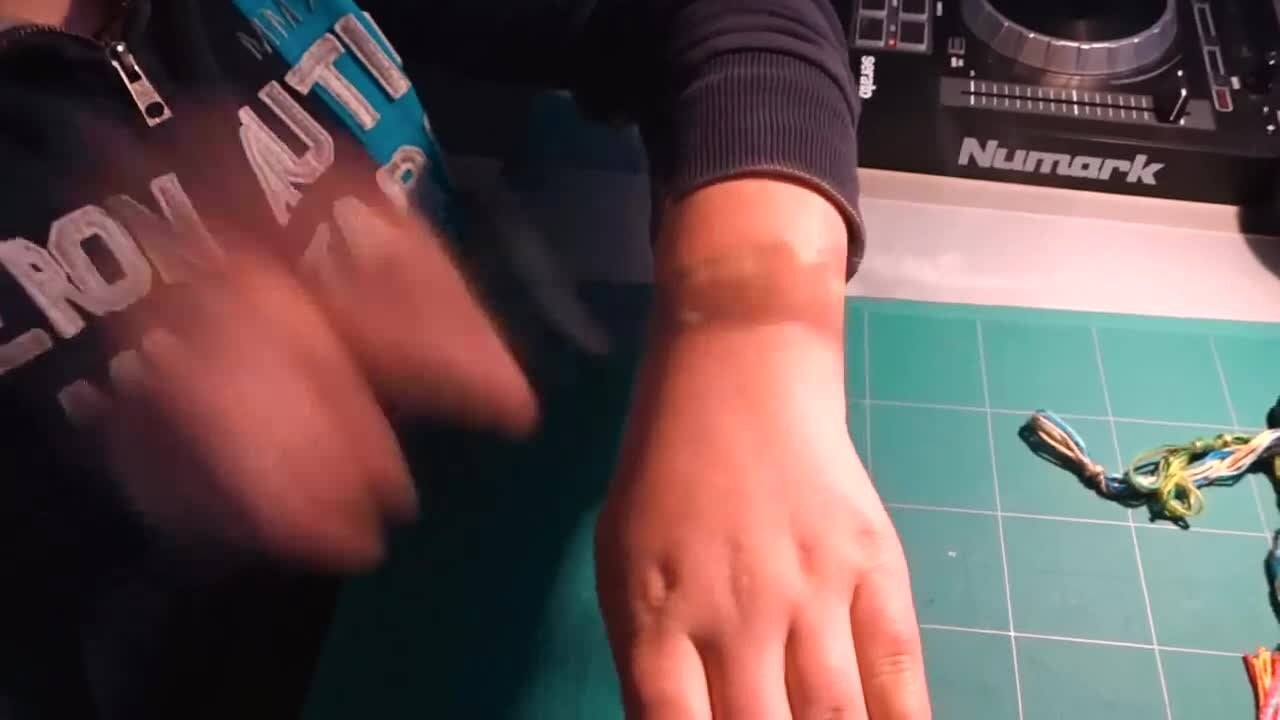 A person wearing a dark sweatshirt sits at a green cutting mat, rubbing their wrist. Colorful embroidery threads and part of a Numark DJ controller are also visible on the table.