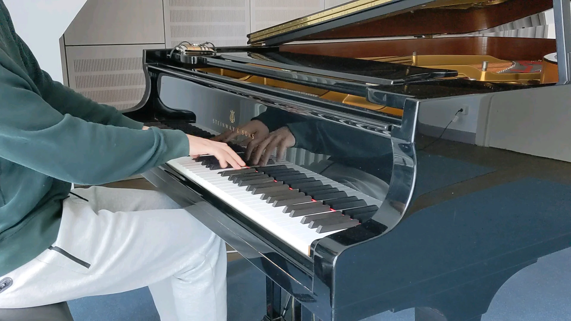 Person wearing a green sweatshirt and light pants is playing a black Steinway & Sons grand piano, with sheet music visible on the top. Only their upper body and arms are in view.