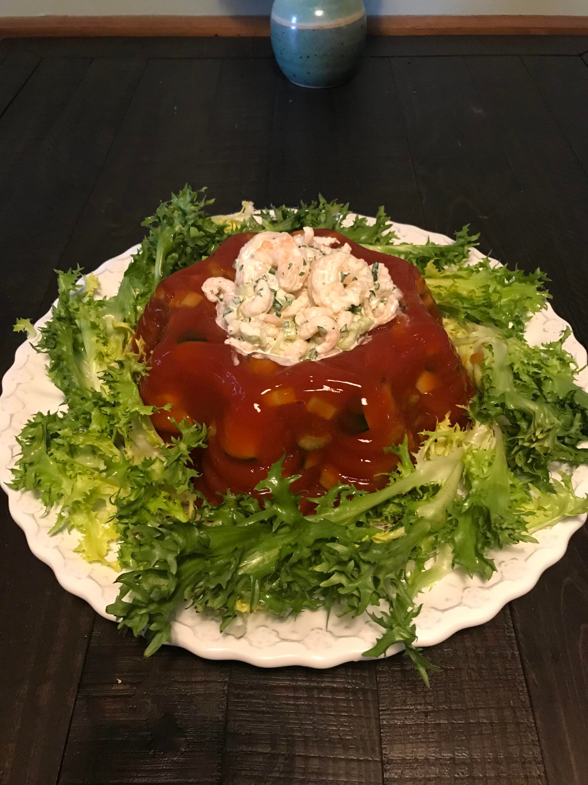 A tomato aspic with vegetables is garnished with fresh greens and topped with a creamy shrimp salad, served on a white plate on a dark table.