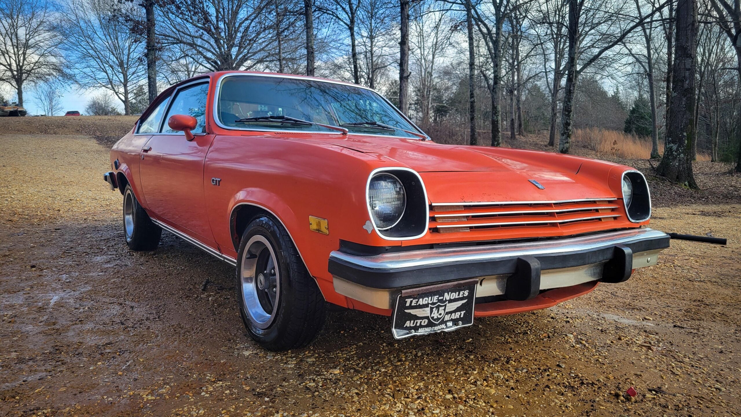 An orange vintage car with a black roof is parked on a gravel road, surrounded by leafless trees. The license plate reads “Teague-Noles Auto Mart.” The sky is partly cloudy.
