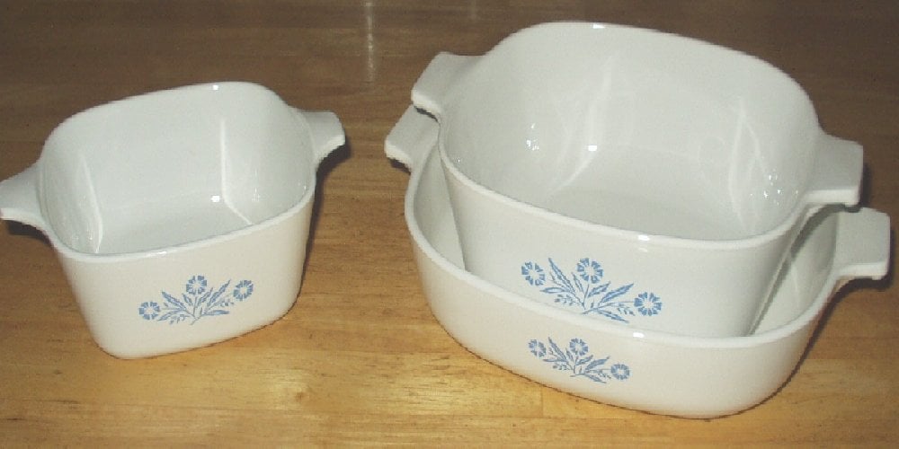 Three white ceramic casserole dishes with blue floral patterns, two stacked on the right and one smaller dish on the left, are placed on a wooden surface.