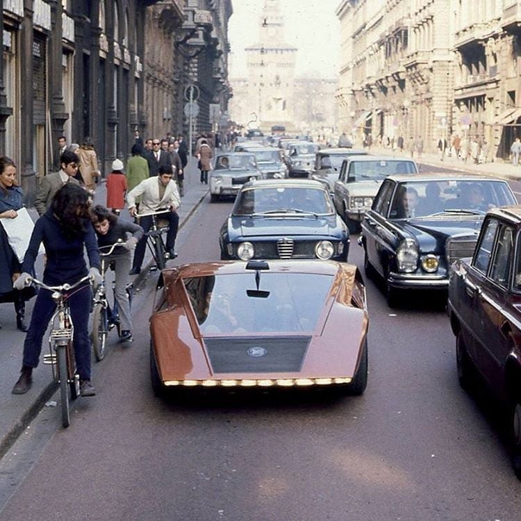 A futuristic, low-slung orange car drives down a busy city street with classic cars, cyclists, and pedestrians nearby. Tall buildings line both sides of the road.