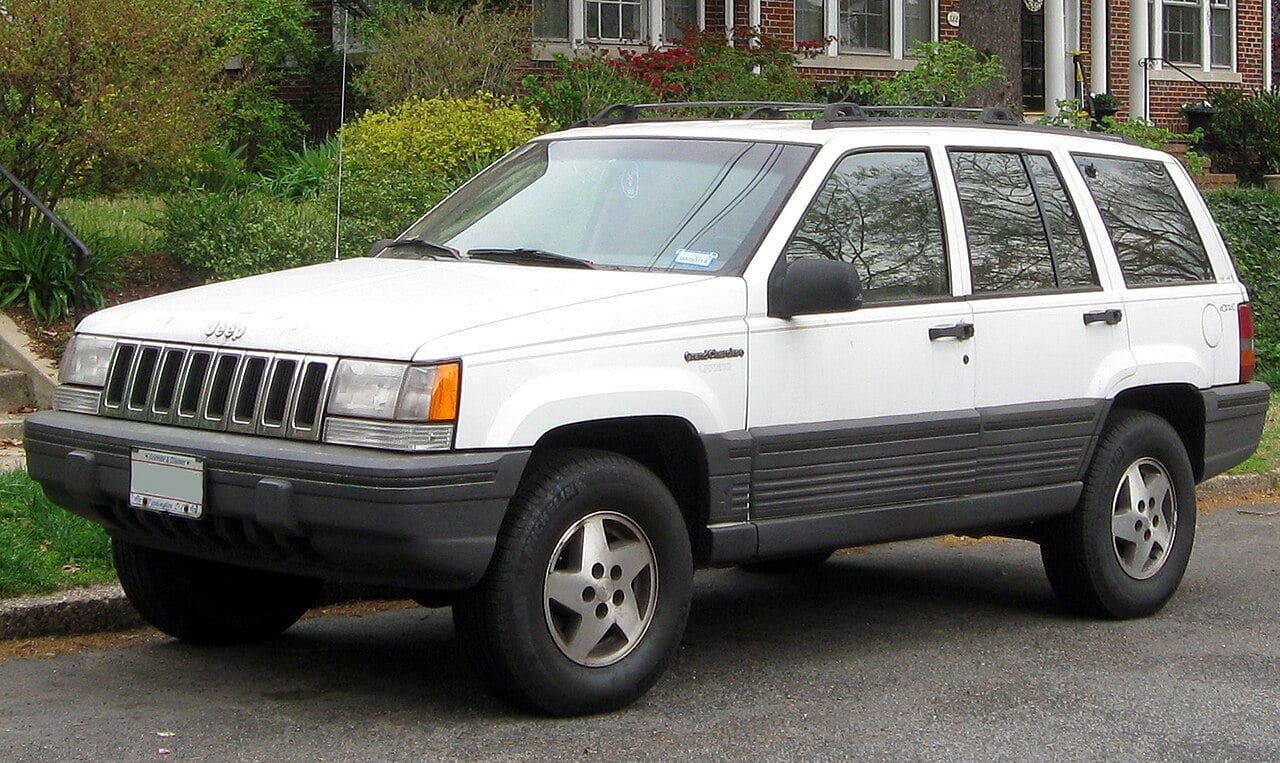 A white Jeep Grand Cherokee SUV is parked on the street in a residential neighborhood, with a brick house and green bushes in the background.