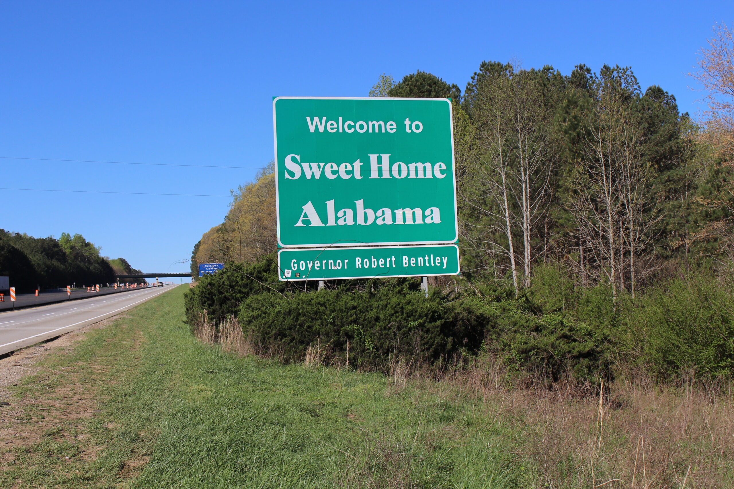 A green roadside sign reads “Welcome to Sweet Home Alabama, Governor Robert Bentley” beside a highway, with trees and blue sky in the background.