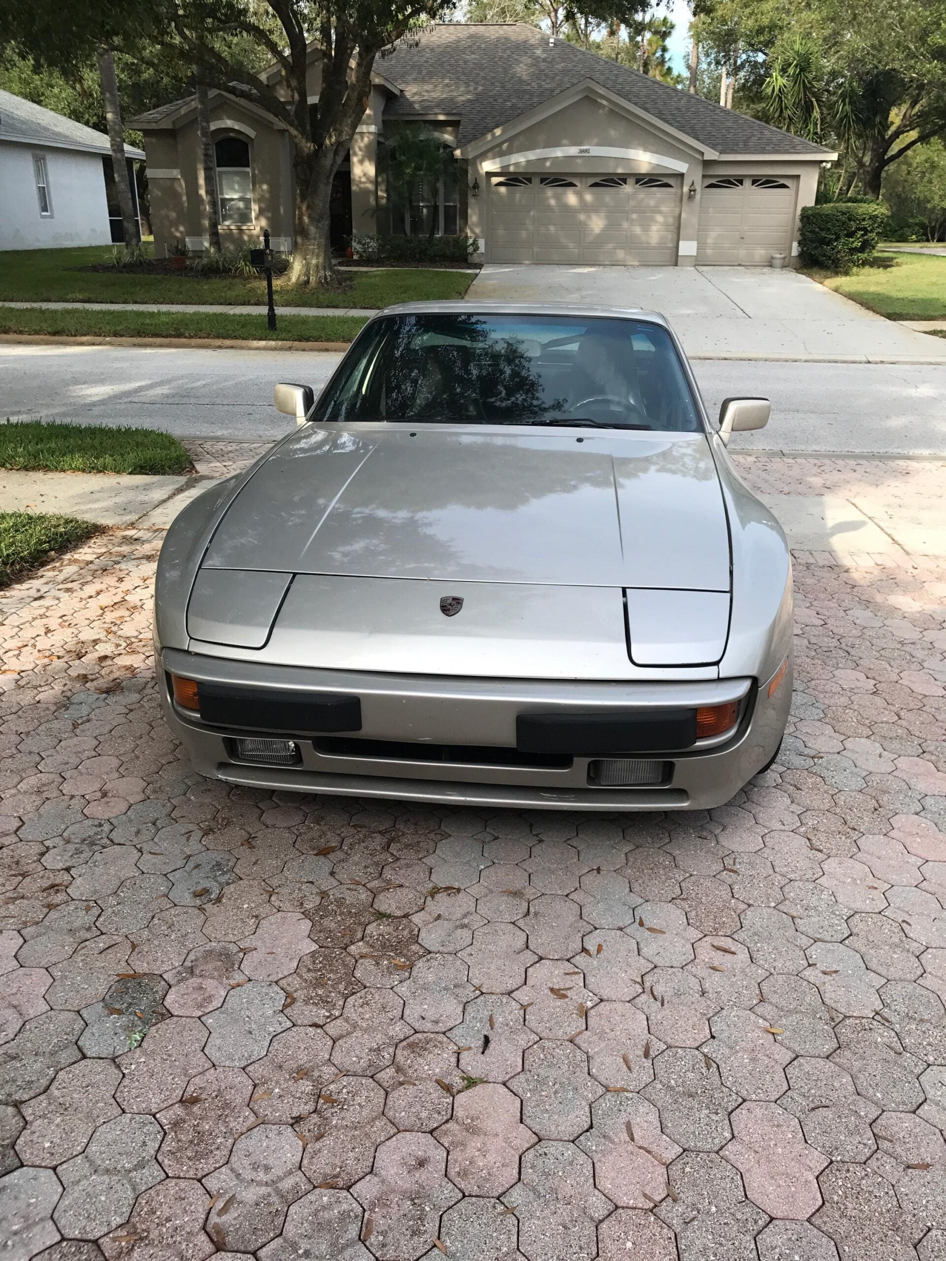 A silver Porsche sports car is parked on a brick driveway in front of a suburban house with a driveway and trees in the background.