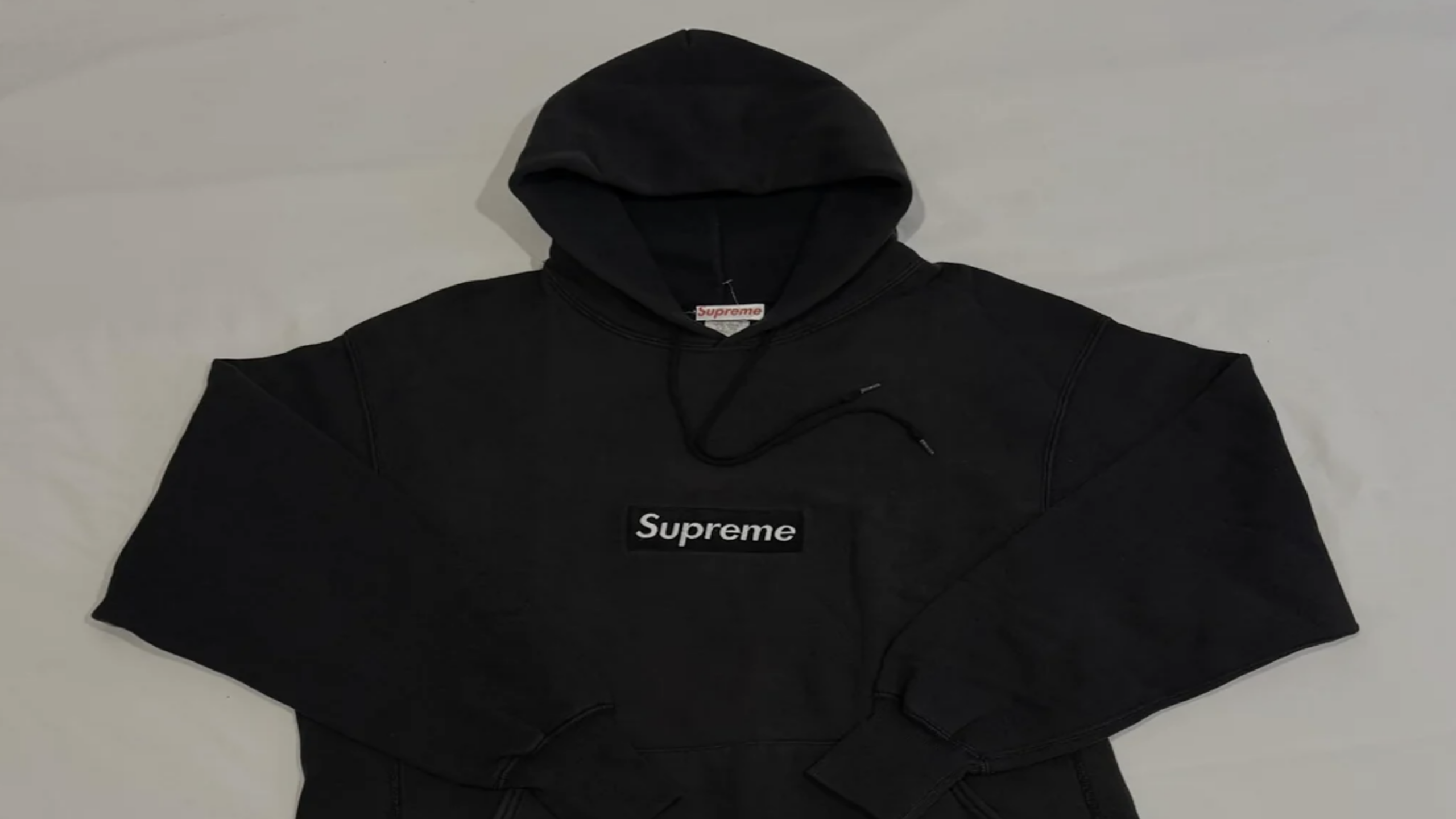 A black Supreme hoodie with a hood, front pocket, and the word “Supreme” embroidered in white on a black rectangular patch on the chest, laid flat on a light surface.
