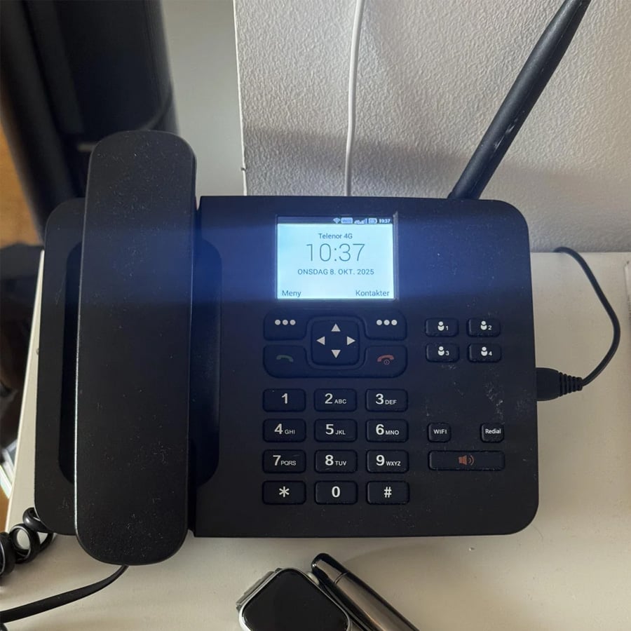 A black desk phone with a screen displaying the time “10:37” and date “ONS DAG 8. OKT. 2025” sits on a white surface. The phone’s receiver rests on the left side, and an antenna is attached on the right.