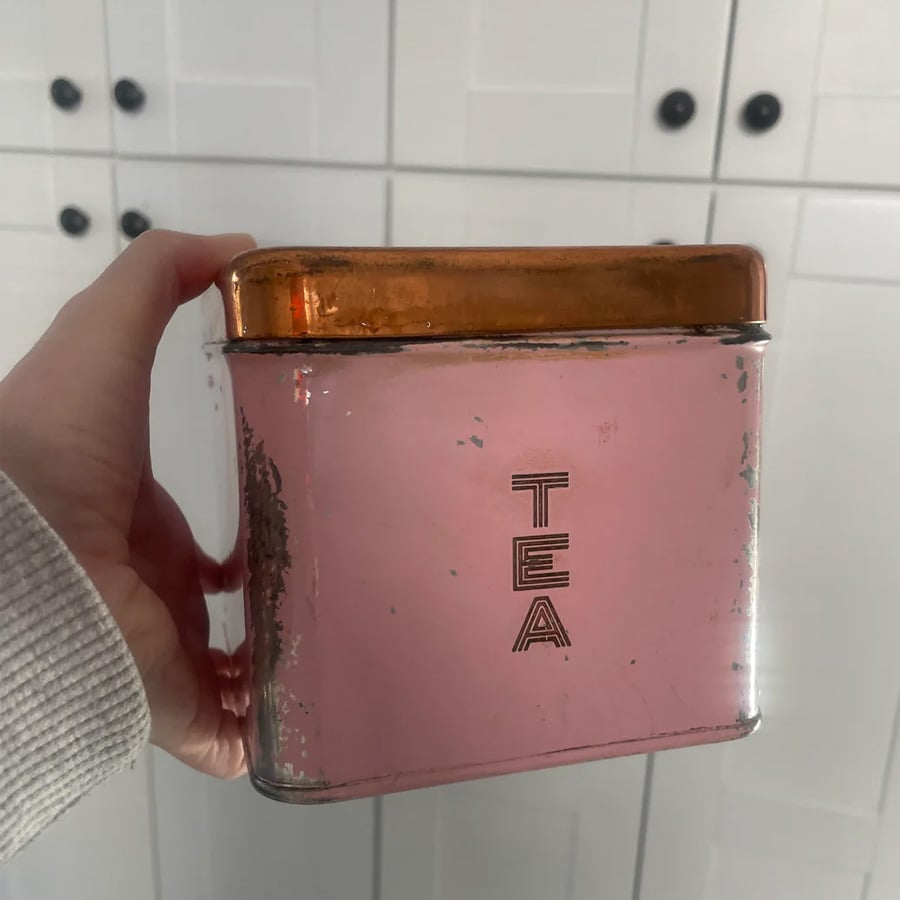 A hand holds a pink vintage tin with a gold lid labeled “TEA” in capital letters. The tin appears slightly worn. White cabinets with black knobs are in the background.