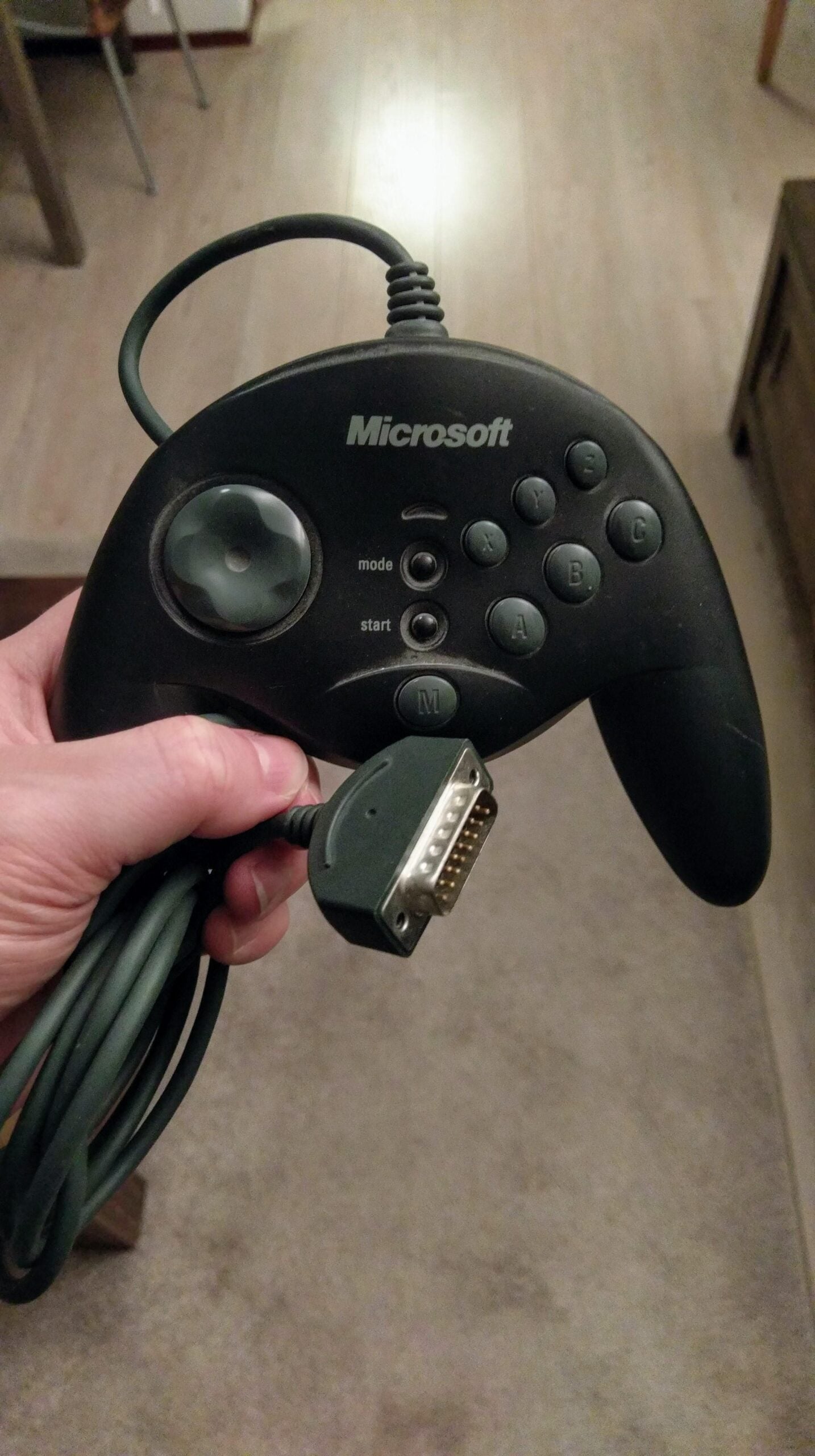 A hand holds a vintage black Microsoft game controller with a joystick, several buttons, and a serial connector plug, against a background of a wooden floor and carpet.