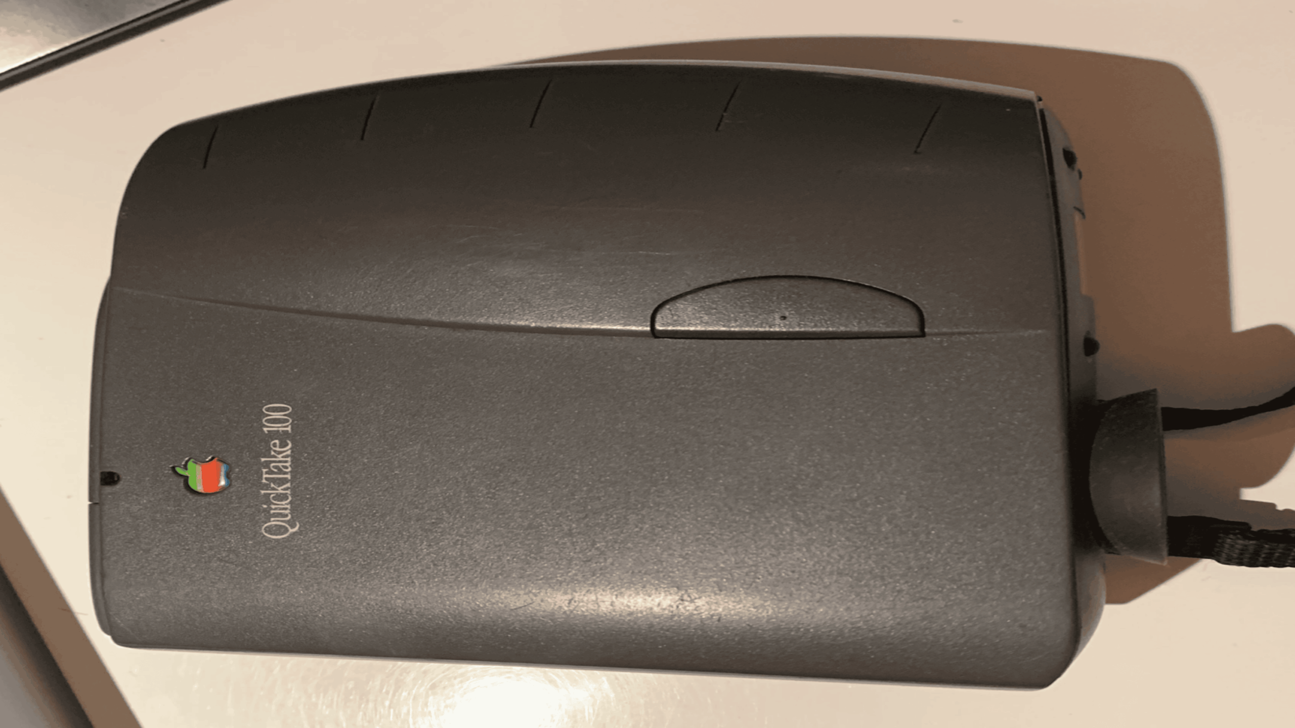A gray Apple QuickTake 100 digital camera rests on a white surface, showing its top side with the Apple logo and product name visible.