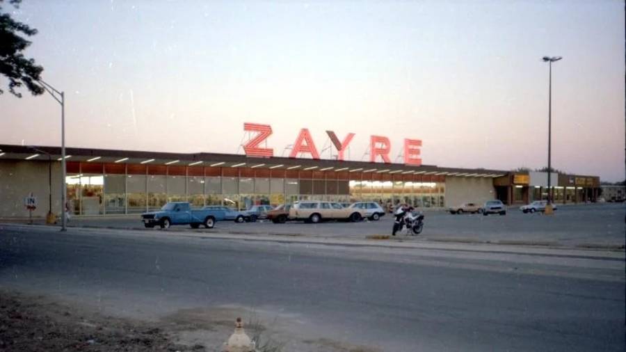 A vintage Zayre store with large red letters on the roof, cars parked in front, and a mostly empty parking lot at dusk.