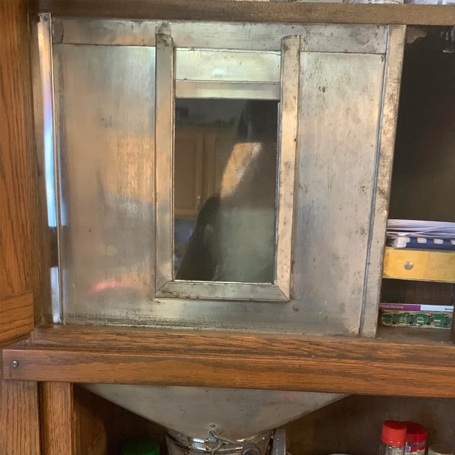 A metal breadbox with a glass window in the center sits on a wooden shelf, with part of a funnel and some jars visible below, and notebooks or papers stacked to the right.
