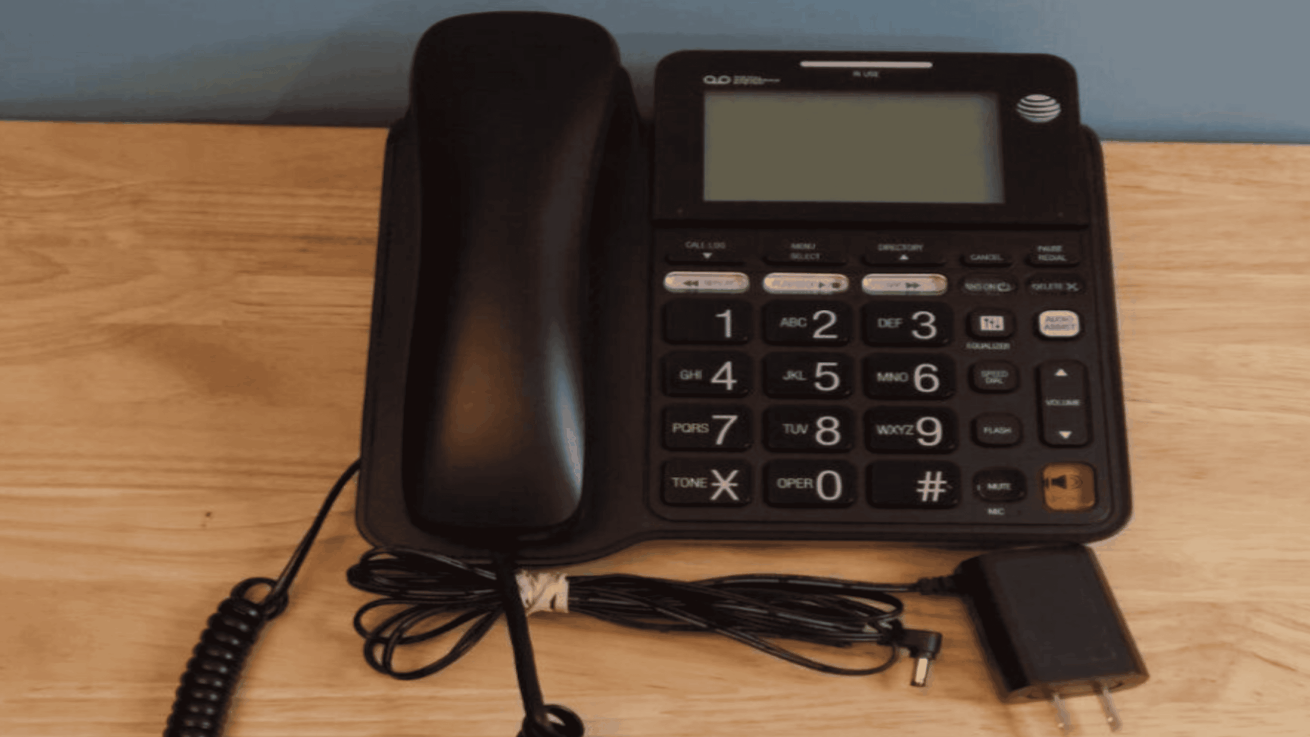 A black AT&T corded landline phone with large buttons sits on a wooden surface. The coiled cord and power adapter are placed in front of the phone.