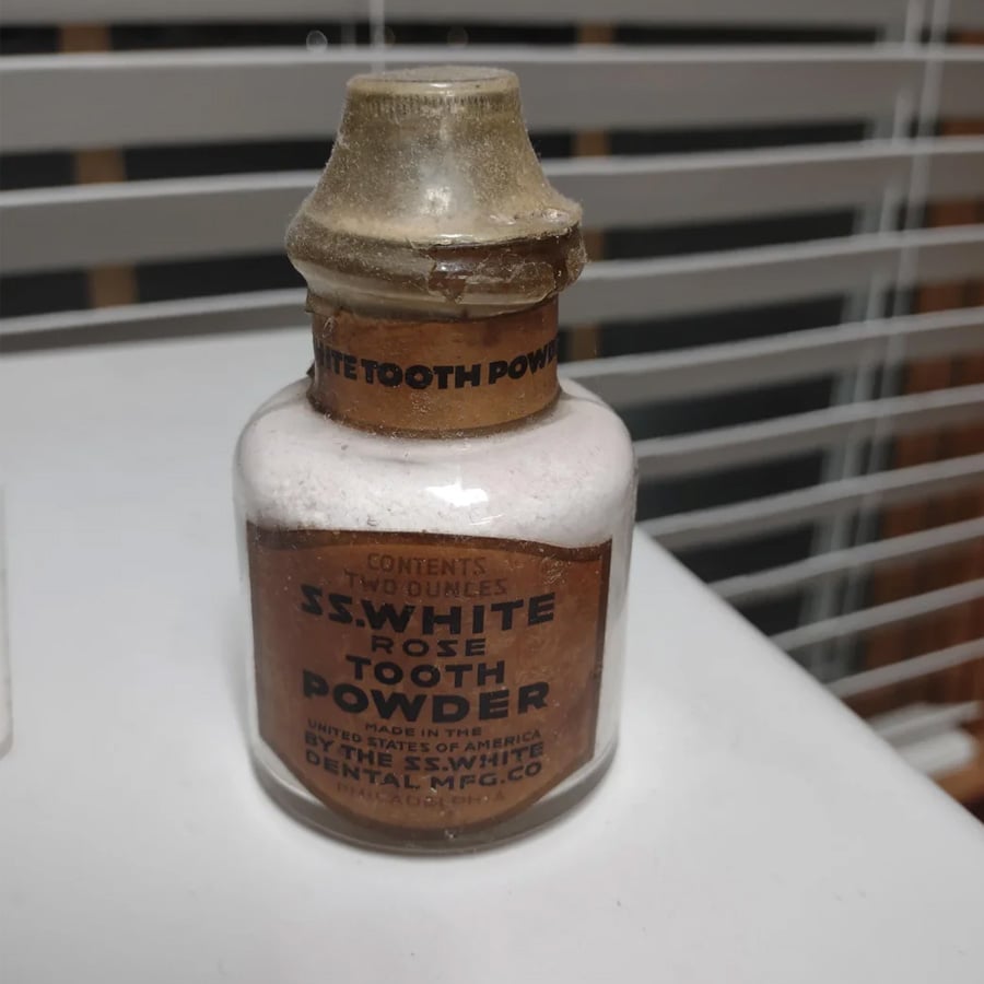A vintage glass bottle labeled “S.S. White Rose Tooth Powder” with a cork and paper seal, sitting on a white surface near window blinds.